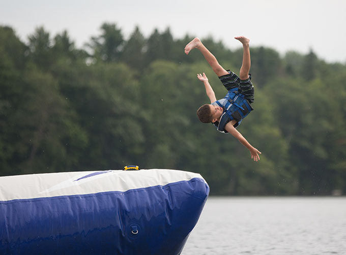 A camper wearing a lifejacket jumps upside down off the water trampoline into Spider Lake at North Star Camp for Boys.