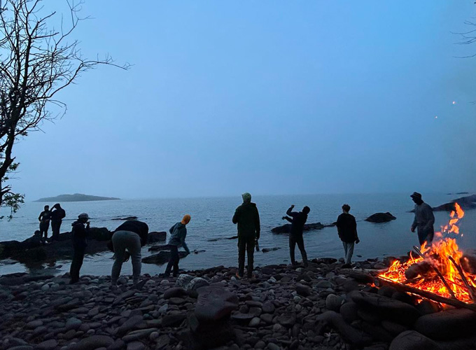 A group of campers at North Star Camp for Boys skip rocks into the lake, while standing on the pebbly shore. A bonfire is burning on the shore.