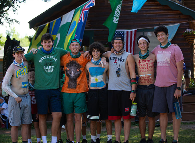 A group of campers and counselors smile with their arms around each other under the international flags at North Star Camp for Boys.