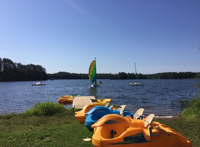 Image of Fun Bug boats sitting along the shores of Spider Lake, and sailboats in the distance in the lake at North Star Camp for Boys.