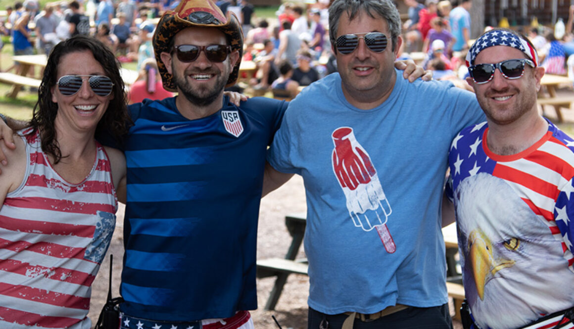 North Star Camp for Boys directors Robyn, Andy, Dan, and Brian wear fourth of July clothing and smile and laugh with their arms around each other during a picnic celebration.