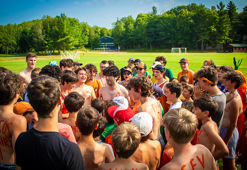 A group of campers huddle around in a circle wearing orange bandanas and body paint while creating a game plan for an activity at North Star Camp for Boys.