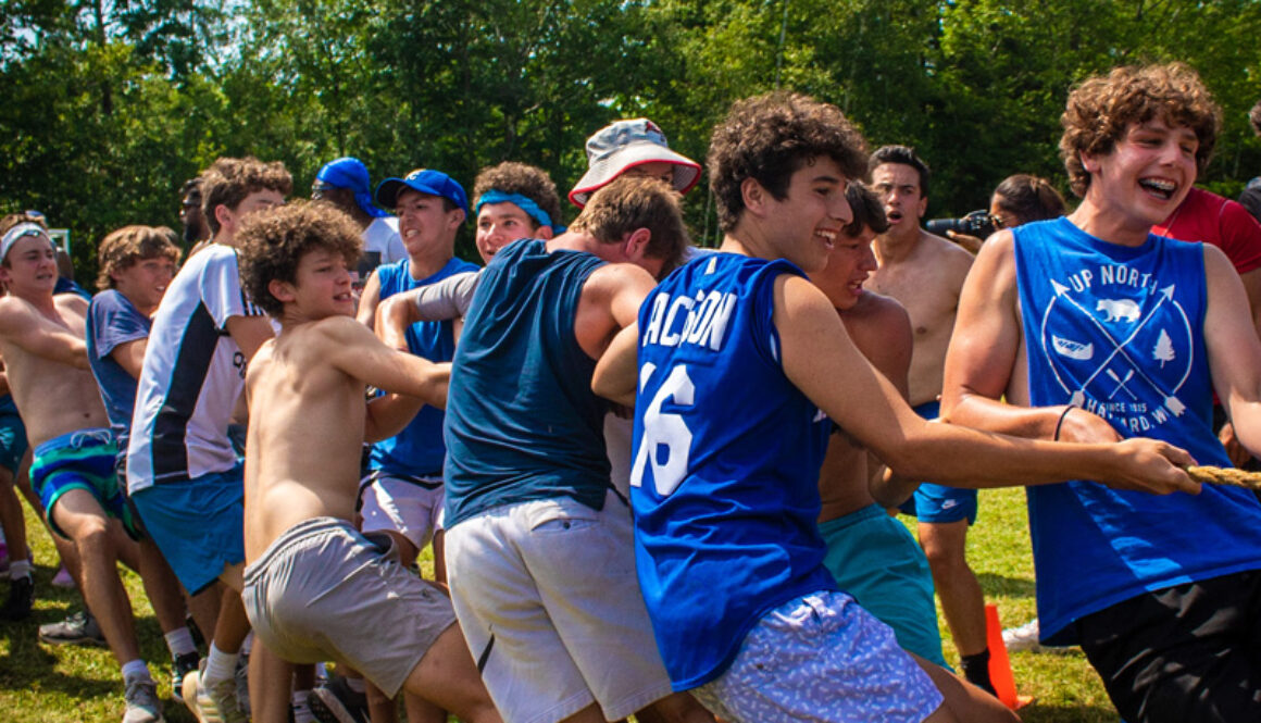 Campers wearing blue and grey smile and laugh while grouped around a rope, tugging and pulling, in an attempt to win a game of tug of war at North Star Camp for Boys.