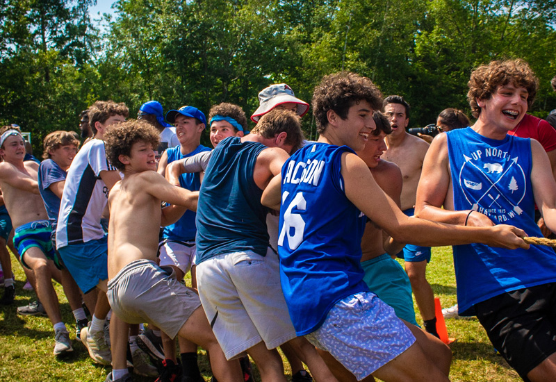 Campers wearing blue and grey smile and laugh while grouped around a rope, tugging and pulling, in an attempt to win a game of tug of war at North Star Camp for Boys.