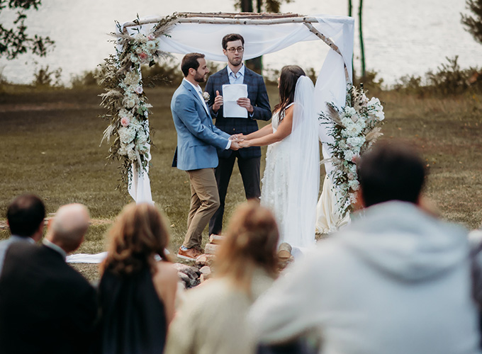 A couple wearing wedding attire - the bride in her white dress and long veil, the groom in a blue suit jacket and khaki pants- hold hands and listen to their officiant while standing under a wood and floral awning during their wedding ceremony at the North Star Camp for Boys facility.