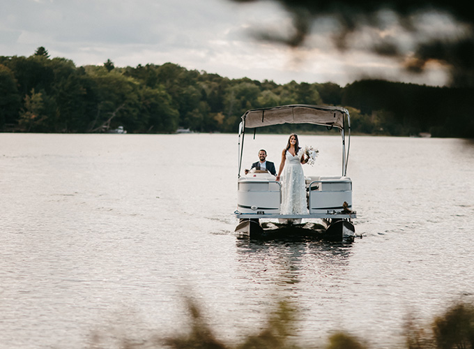 A bride wearing her wedding dress stands on a pontoon boat, being driven by her brother down Spider Lake, and smiles while holding her bouquet on her wedding day at the North Star Camp for Boys facility.