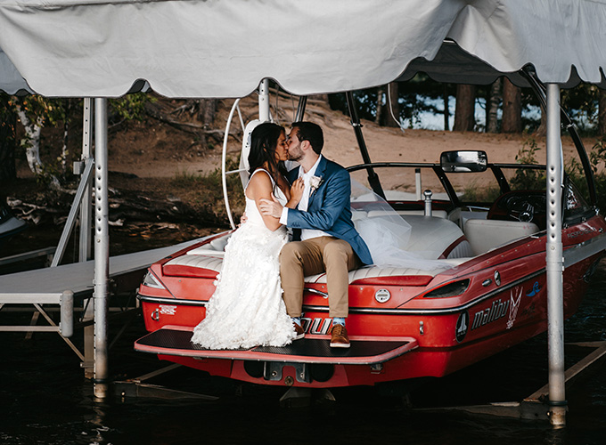 A newlywed couple, the bride wearing her wedding dress, and the groom wearing a blue suit jacket and khaki pants, kiss while sitting on a red speedboat in Spider Lake at their wedding at the North Star Camp for Boys facility.