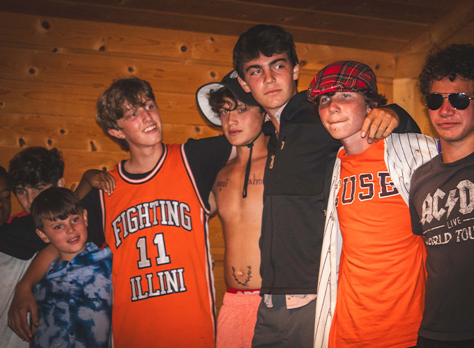 A group of six campers, two wearing orange, stand with their arms around each other while participating in a theater play at North Star Camp for Boys.