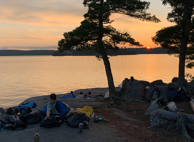 Campers lay in sleeping bags on a rock formation to the side of the lake watching the sunset at Quetico Provincial Park in Ontario, Canada during "The Canadian" North Star Camp for Boys wilderness trip.