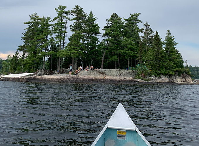 A canoe in the river at Quetico Provincial Park points to the shore where campers are seated on a rock formation during a wilderness trip at North Star Camp for Boys.