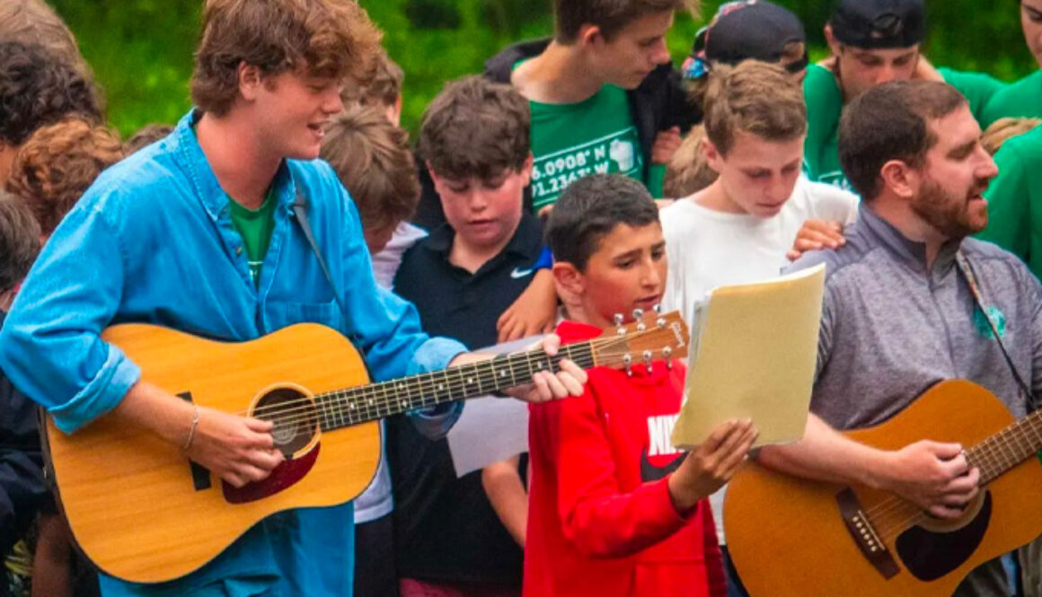 A group of campers stand with their arms around each other singing a song while two staff members play the guitar during a Friday Night Service at North Star Camp for Boys.