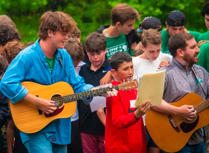 A group of campers stand with their arms around each other singing a song while two staff members play the guitar during a Friday Night Service at North Star Camp for Boys.