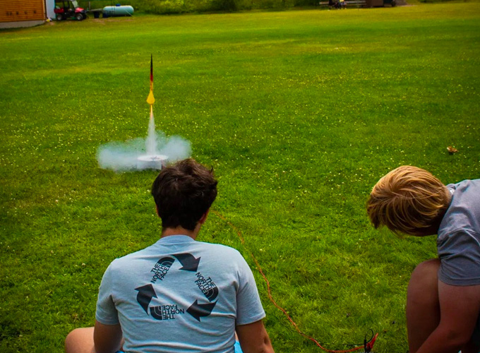 Two campers watch on as a red and yellow rocket begins to lift off the ground, smoke surrounding it, during a rocketry activity at North Star Camp for Boys.