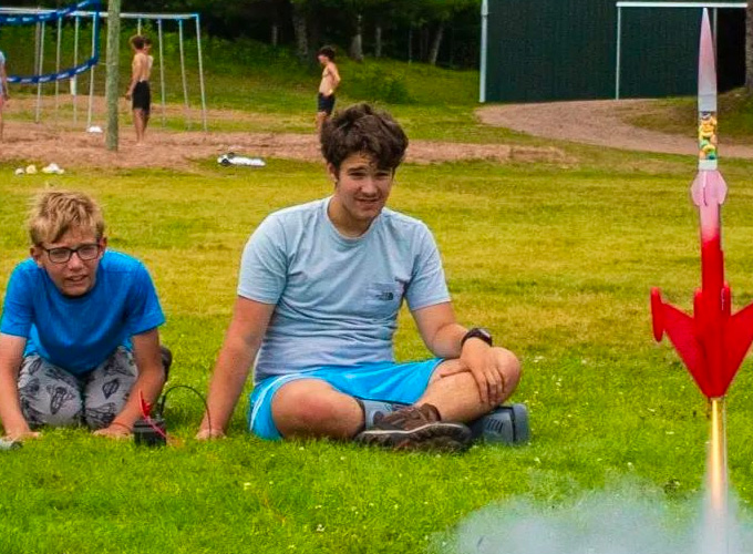 A camper and staff member watch as a red and pink painted rocket begins to blast off the ground, leaving a trace of smoke during a rocketry activity at North Star Camp for Boys.