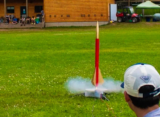 A red and gold painted rocket leaves a small circle of smoke as it begins to lift off the ground, as a camper wearing a baseball hat watches on during an activity at North Star Camp for Boys.