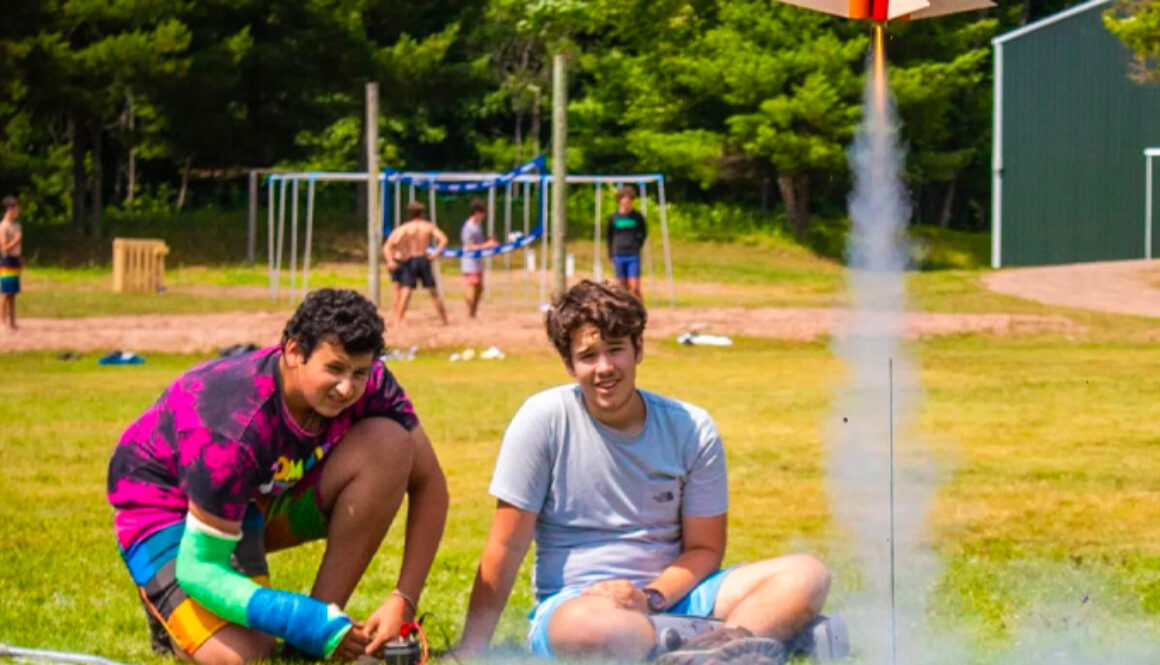 Two staff members at North Star Camp for Boys watch a rocket take off, leaving a trace of smoke, during a rocketry activity at North Star Camp for Boys.