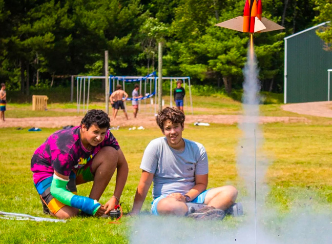 Two staff members at North Star Camp for Boys watch a rocket take off, leaving a trace of smoke, during a rocketry activity at North Star Camp for Boys.