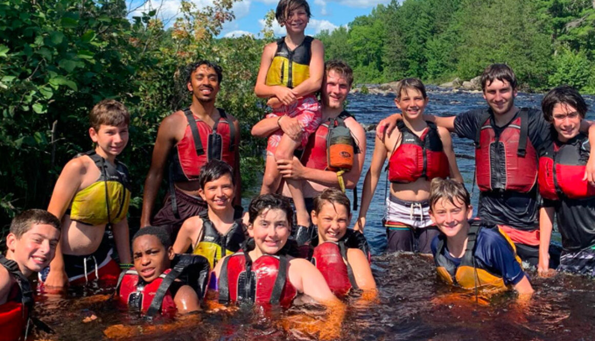 Campers and trip leaders wear life jackets and smile while swimming in the Flambeau River during a wilderness trip at North Star Camp for Boys.