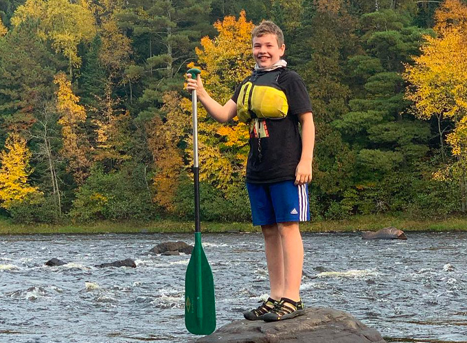 A camper wearing a life jacket smiles while standing on a rock formation on the Flambeau River holding a canoe ore during a wilderness trip at North Star Camp for Boys.