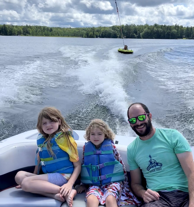 A dad smiles with his two daughters as they sit on a boat and another person goes tubing during family camp at North Star Camp for Boys.