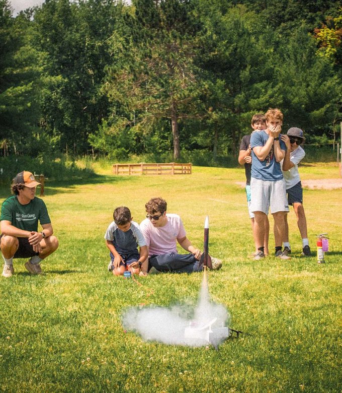 Campers watch a model rocket launch on the field.